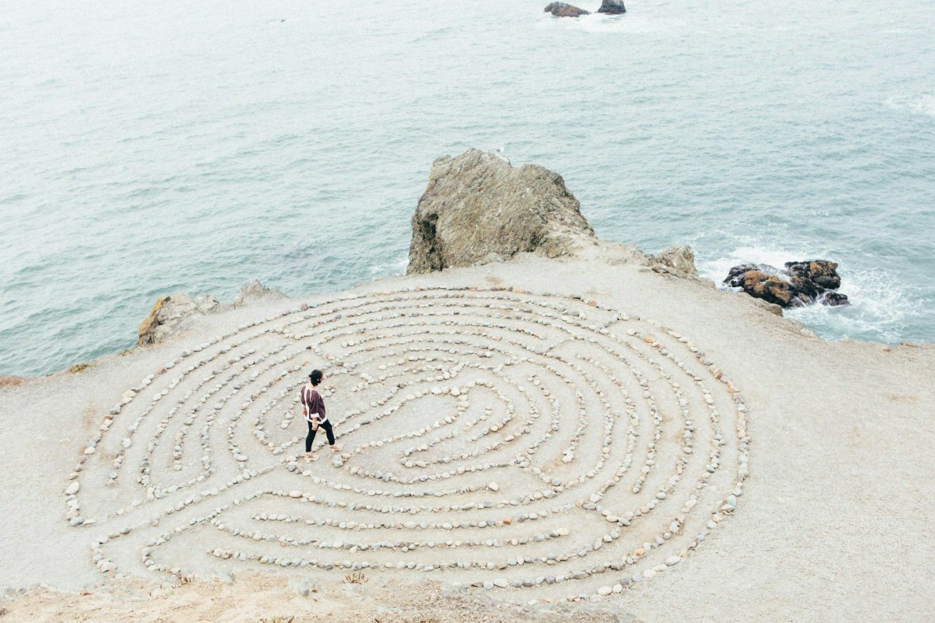 Weg zur Klarheit – Coaching am Meer Vogelperspektive auf einen Strand mit Felsen, im Sand ein gezeichnetes Labyrinth, in das ein Mensch eintritt – Symbol für Orientierung, Refl